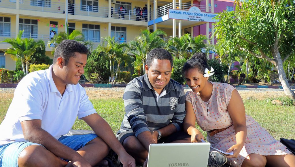 三名学生坐在斐济大学校园的一栋大楼外。